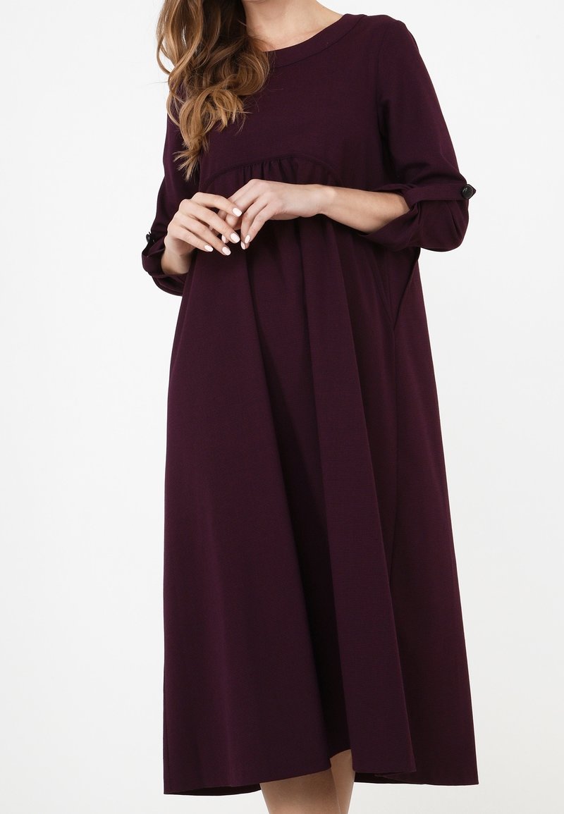 Robe violette foncée à coupe ample, manches longues, décolleté rond, détails froncés au niveau de la poitrine et accents en boutons sur les manches.