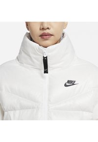 Balta pūkinė striukė su aukštu apykakle, matomu užtrauktuku ir juodu Nike logotipu ant krūtinės, pasižyminti siuvinėtu tekstūru ir bespalviu dizainu.