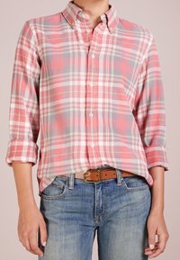 Röd och rosa rutig skjorta med knapp-ner krage, långa ärmar uppvikta, och en liten logotyp på bröstet; kombinerad med blå jeans och ett brunt bälte.