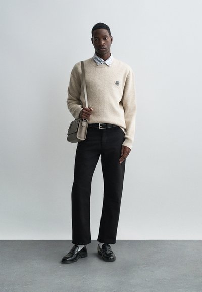 Bēša vilnas džemperis ar logo, blūze ar apkakli, melni plaši bikses, melnas kurpes un pelēka soma. Tekstūri audums un brīva piegriezuma stils.