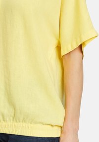 Żółta lniana bluzka o luźnym kroju, z krótkimi rękawami i elastycznym dołem. Tekstura wydaje się miękka i lekka.