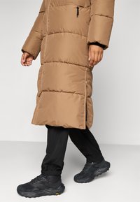 Brun polstret lang jakke med glidelås og sidelommer, brukt over svarte smale bukser og svarte teksturerte joggesko.