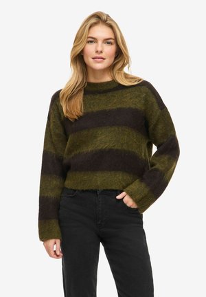 Nainen pukeutuneena vihreään ja mustaan leveäraidalliseen pörröiseen neulepaitaan ja mustiin housuihin, poseeraa yhdellä kädellä taskussa valkoista taustaa vasten.