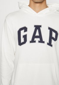 Felpa bianca con tasca frontale, caratterizzata da lettering "GAP" in blu navy in un font spesso e ricamato. Tessuto morbido con cappuccio e vestibilità casual.