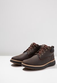 Bruine leren enkelboots met een rubberen zool, voorzien van gouden oogjes, bruine veters en een gestructureerd ontwerp op de zool voor grip.