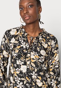 Blusa preta com flores amarelas e brancas, decote franzido e lista de botões. Tecido leve com padrão de bolinhas.