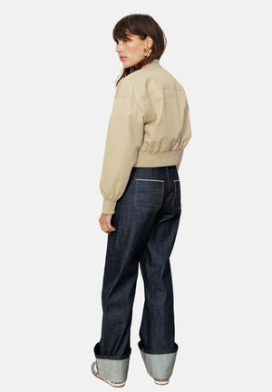 Femme portant une veste beige courte, un jean large bleu foncé avec ourlets retroussés, et des chaussures imprimé léopard, de profil sur fond blanc.