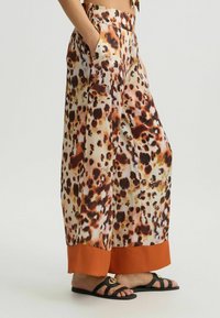 Luipaardprint wijdbeenbroek van zacht materiaal, met een oranje zoom, zijzakken en een losse pasvorm voor comfort. Er worden platte sandalen gedragen.