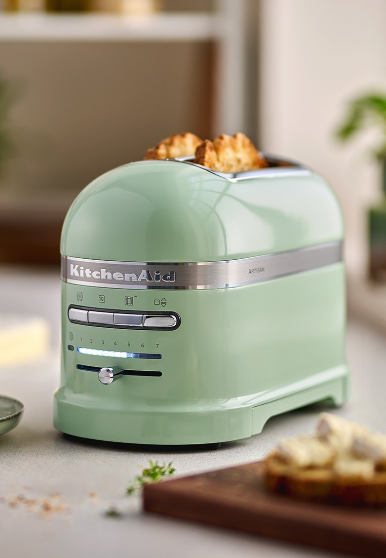 Grille-pain KitchenAid vert au design arrondi, avec des accents en acier inoxydable et sept niveaux de brunissage ; il dispose d'un tiroir à miettes à l'avant et de commandes éclairées.