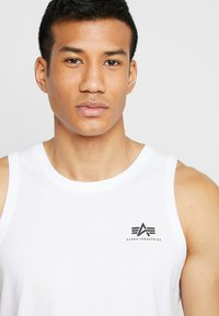 Bijela pamučna majica bez rukava s crnim Alpha Industries logotipom na lijevom prsnom dijelu. Glatka tekstura, okrugli izrezi i bez rukava.