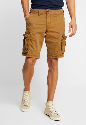 Homme portant un short cargo marron avec poches à rabat et des baskets montantes beiges, debout devant un fond clair uni.