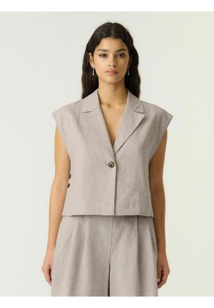 Jeune femme portant un blazer beige sans manches avec un seul bouton et un pantalon taille haute assorti, debout devant un fond clair uni.
