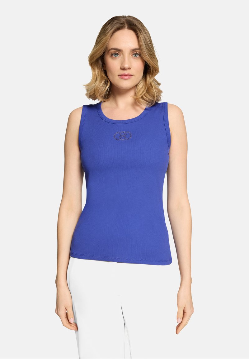 Jeune femme aux cheveux blonds portant un haut bleu sans manches et un pantalon blanc, debout devant un fond clair uni.