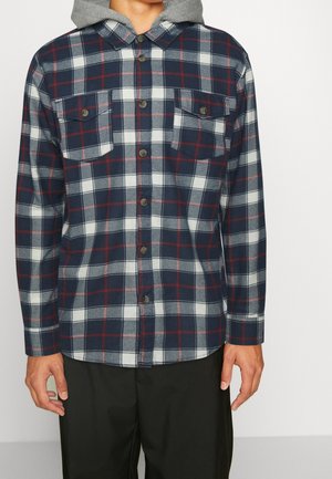 Man draagt een marineblauw, wit en rood geruit overhemd met knopen, met twee borstzakken en een grijze capuchon eronder.