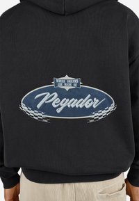 Sort hættebluse med et cirkulært blåt grafik, der har teksten "Pegador" og sætningen "Where dreams are made of." Tjekkede detaljer pryder designet.