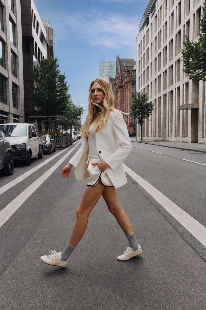 Femme aux longs cheveux blonds portant un blazer blanc, un short, des chaussettes grises et des chaussures blanches marchant dans une rue urbaine vide entre des bâtiments modernes.