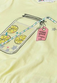 Žluté tričko s grafikou zavařovací sklenice se plátky citronu, pruhovaným brčkem a růžovým štítkem s nápisem "DOMÁCÍ limonáda." Ozdobené flitry.