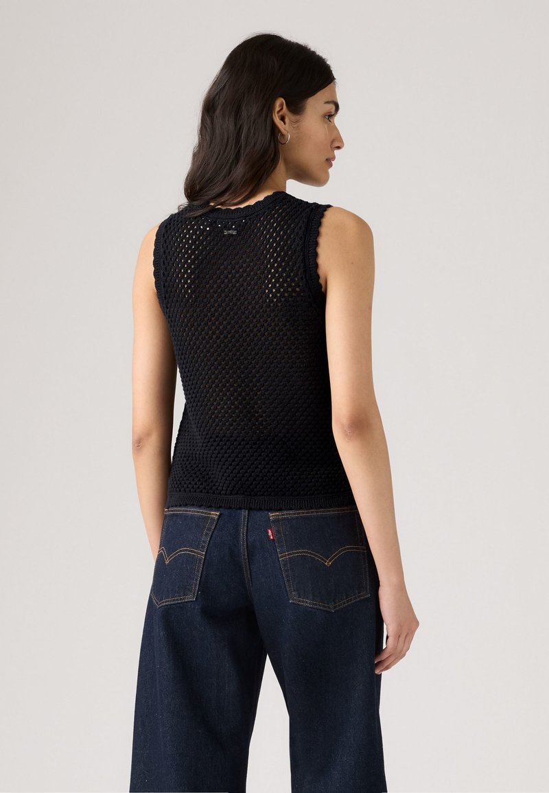 Kobieta z ciemnymi włosami, stojąca bokiem, ubrana w bezrękawnik z czarnej dzianiny i ciemnoniebieskie dżinsy o szerokich nogawkach, na jednolitym tle.