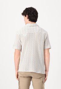 Polo de manga corta en beige claro con un patrón texturizado. Cuenta con un cuello y un detalle de bolsillo trasero. La tela parece transpirable.