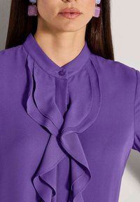 Frau trägt eine lila Bluse mit Rüschendetails auf der Vorderseite und passenden lila quadratischen Ohrringen, gezeigt von Hals bis Brust.