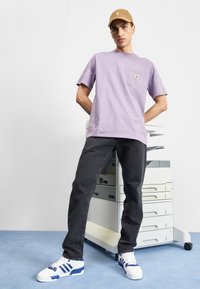 Lyselilla bomull t-skjorte med brystlomme, kombinert med mørke jeans og hvite sneakers med blå striper. Bakgrunn: kontorprinter.