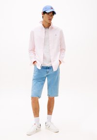 Lichtroze overhemd met knoopjes over een gestreept grijs-wit t-shirt, gecombineerd met lichtblauwe denim shorts en witte sneakers. Blauwe pet op het hoofd.