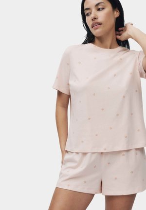 Kvinde iført lyserød top med korte ærmer og matchende shorts med små ferskenmønstre, poserer med den ene hånd bag nakken.