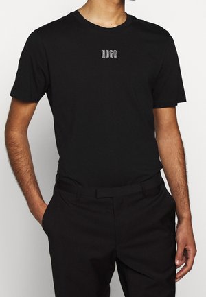 Mies, jolla on musta, istuva, lyhythihainen T-paita, jossa pieni valkoinen "HUGO"-logo rinnassa, ja mustat housut, kädet osittain taskuissa.