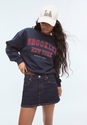 Felpa blu navy con la scritta "BROOKLYN NEW YORK" in rosso, abbinata a una mini gonna in denim scuro. Un cappellino color crema mostra la scritta "LA - SUNNY DAYS."