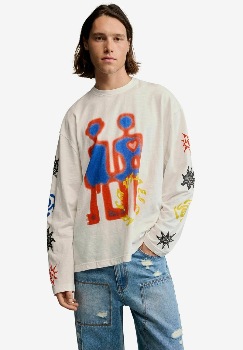 Junger Mann mit schulterlangem Haar, der ein weißes Langarmshirt mit bunten abstrakten Figuren und Sonnendesigns trägt, kombiniert mit hellen Jeans.