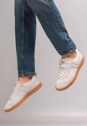 Beine in blauen Jeans, die weiße Sneakers mit braunen Sohlen tragen, in der Luft vor einem einfarbigen hellen Hintergrund.