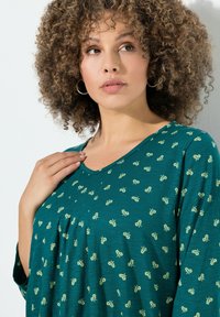 Turfgroene top met korte mouwen, met een bloemenpatroon in lichtgroen, een V-hals en geplooide details aan de voorkant. Zachte stoftextuur.