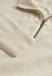 Vaaleanbeige vetoketjupusakka, jossa on teksturoitu ribbikuvio, korkea kaulus ja metallivetoketju. Pehmeä materiaali, jossa on hienovarainen kiilto ja lämpö.