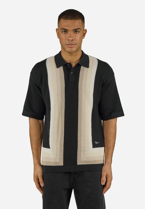 Mann trägt ein schwarzes kurzärmeliges, gestricktes Poloshirt mit beigen und weißen vertikalen Streifen und steht vor einem schlichten Hintergrund.