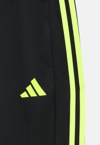 Pantalones deportivos negros con rayas laterales amarillas brillantes y un logo de Adidas en la parte inferior de la pierna izquierda. Hechos de tela texturizada.
