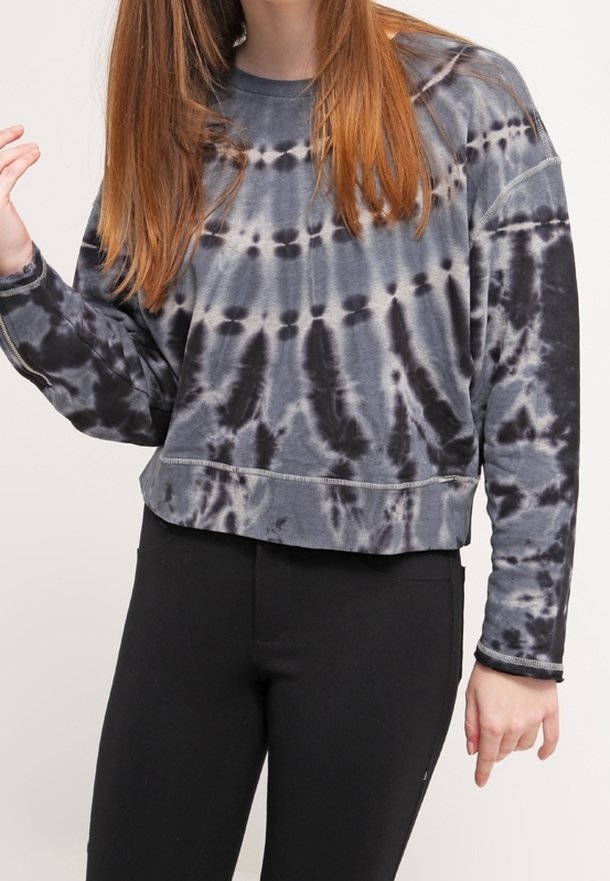 Jeune personne portant un sweat court tie-dye noir et gris à manches longues et un pantalon noir, avec des cheveux raides châtain roux.