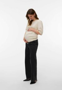 Femme enceinte en pull beige et jean noir tenant doucement son ventre, souriant et regardant vers le bas sur un fond blanc uni.