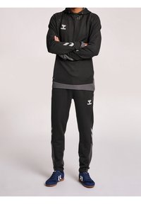 Haut de hoodie de sport noir avec une partie supérieure texturée, accents gris et logo ; associé à un pantalon noir avec des bandes latérales et des chaussures bleues.