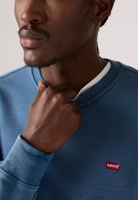 De blauwe sweatshirt van Levi's heeft een ronde hals, ribgebreide manchetten en een klein rood logo-patch. De stof lijkt zacht en textuur te hebben.