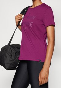 Fialové bavlněné tričko s krátkými rukávy, zdobené texturovaným potiskem "PASSION PURPOSE". Černá sportovní taška se zipem držena v levé ruce.