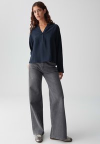 Donna con lunghi capelli ricci che indossa una blusa scura a V blu navy, jeans grigi a gamba larga e sneakers argentati su uno sfondo chiaro e semplice.