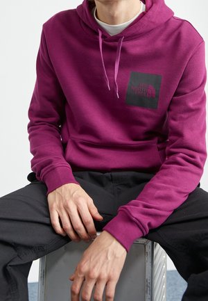 Osoba ubrana w magentową bluzę z kapturem z logo The North Face na piersi, czarne spodnie, siedzi z rękami spoczywającymi na nogach.