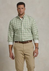 Groene en witte geruite button-down overhemd met lange mouwen, gedragen over een wit shirt; gecombineerd met beige broeken en een bruine riem.