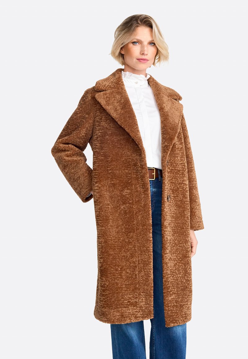 Brauner, strukturierter langer Mantel mit breitem Kragen, vorderem Knopfverschluss und Seitentaschen, kombiniert mit einem weißen Hemd und blauen Jeans.