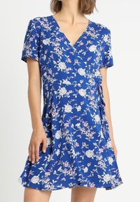 Sininen kukallinen kietaisumekko lyhyillä hihoilla, jossa on valkoisia ja vaaleanpunaisia kukkia. Kevyt kangas, jossa on pehmeä tekstuuri ja vyötäröllä solmittava yksityiskohta.