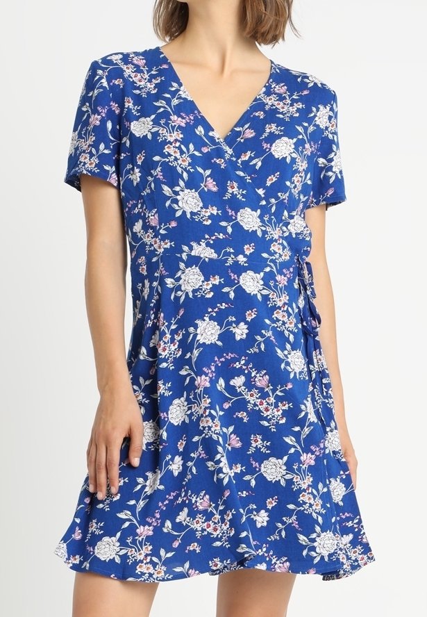 Sininen kukallinen kietaisumekko lyhyillä hihoilla, jossa on valkoisia ja vaaleanpunaisia kukkia. Kevyt kangas, jossa on pehmeä tekstuuri ja vyötäröllä solmittava yksityiskohta.