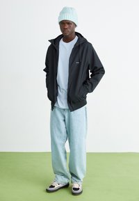 Chaqueta negra con cremallera y capucha, camiseta azul clara, pantalones vaqueros holgados azul claros y zapatillas negras con acentos crema. Con un fondo de suelo verde.
