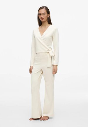 Femme portant un haut cache-cœur couleur crème avec un lien sur le côté et un pantalon large assorti, debout pieds nus sur un fond blanc uni.