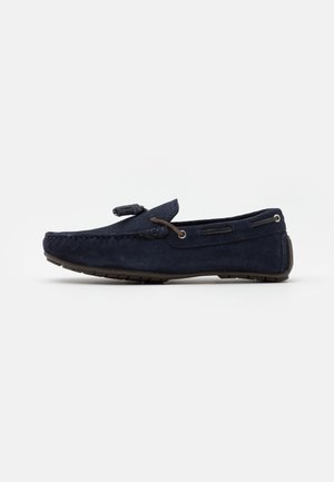 Mocassin en daim bleu marine avec une semelle en caoutchouc basse et un lacet en cuir tressé noué avec un pompon sur le dessus, présenté sur fond blanc.