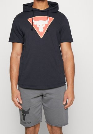 Mann trägt schwarzen Kurzarm-Hoodie mit Stier-Logo und graue Shorts mit passendem Stier-Logo und dem Schriftzug "Project Rock".